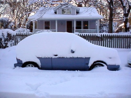 A car completely covered in snow, parked in front of a house with a white picket fence.