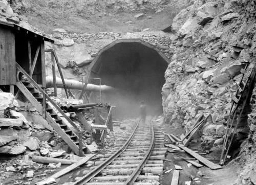 Black and white image of a mine entrance with wooden structures, tracks, and rocky terrain. A figure stands in the distance.