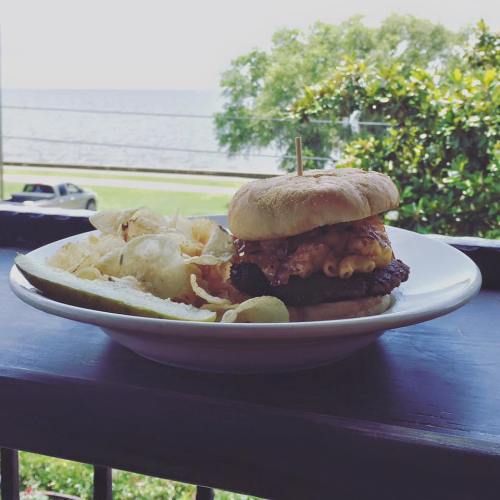 A burger with cheese and macaroni on a plate, served with chips and a pickle, with a scenic view of water in the background.