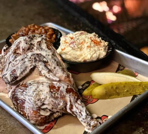 A plate featuring a smoked chicken leg, baked beans, coleslaw, pickles, and lemon wedges on a metal tray.