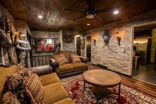 Cozy living room with stone walls, a knight statue, artwork, and a plush sofa on a patterned rug. Warm lighting enhances ambiance.