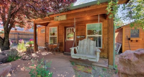 A cozy wooden cottage with a porch swing, surrounded by greenery and colorful flowers.