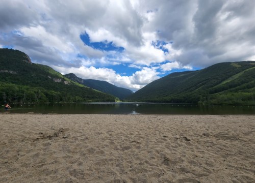A serene lake surrounded by mountains under a partly cloudy sky, with a sandy beach in the foreground.