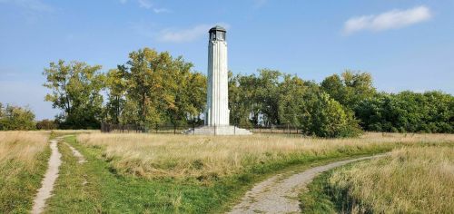 A tall, white monument surrounded by trees and tall grass, with two paths leading towards it under a clear blue sky.