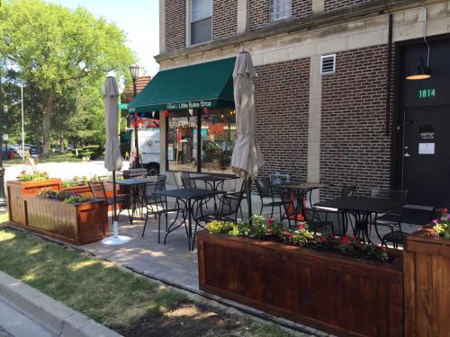 Outdoor seating area with tables and umbrellas in front of a bakery, surrounded by flower planters and a brick building.