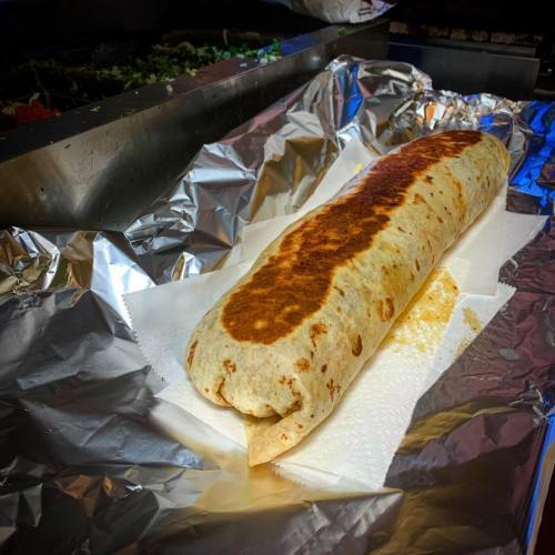 A large, grilled burrito wrapped in foil, resting on a paper towel on a stainless steel surface.