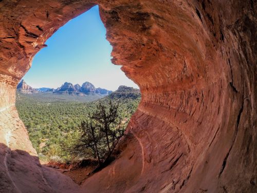 View from a rocky cave opening, showcasing a scenic landscape of mountains and greenery under a clear blue sky.
