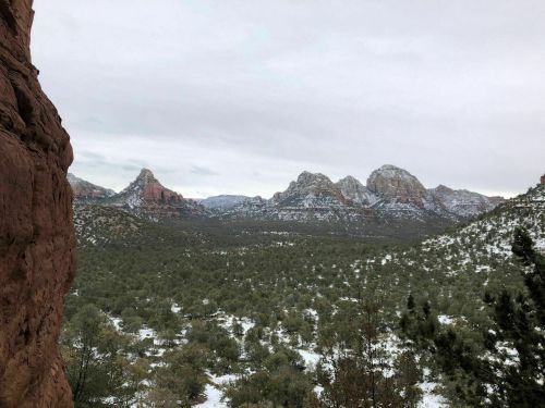 Snow-covered mountains rise above a green valley under a cloudy sky, framed by rocky cliffs in the foreground.