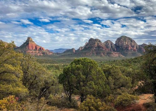A scenic view of red rock formations and green trees under a cloudy sky in a mountainous landscape.