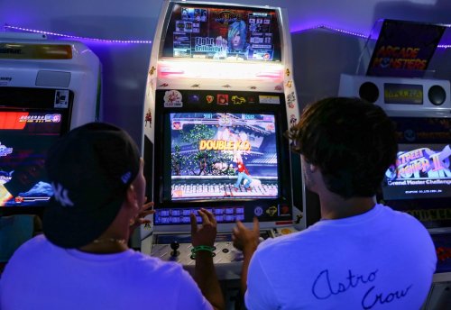 Two players compete at an arcade machine, focused on the screen displaying a fighting game. Neon lights glow in the background.