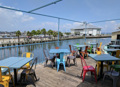 Colorful outdoor seating area with tables and chairs overlooking a marina and water under a clear blue sky.