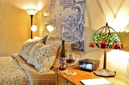 Cozy bedroom scene with a bed, decorative lamp, wine bottles, and a notepad on a wooden nightstand.