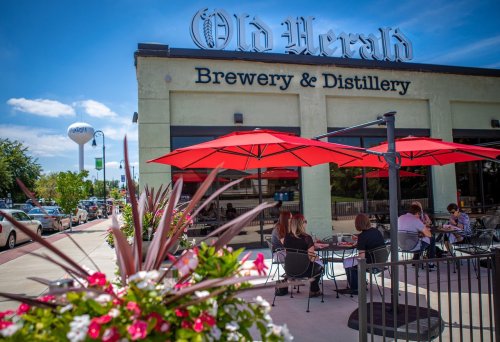 Exterior of Old Herald Brewery & Distillery with red umbrellas, flowers in foreground, and people dining outside.