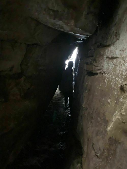 A narrow cave passage with a silhouette of a person standing at the far end, illuminated by light.