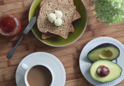 A bowl of toast with butter, sliced avocado, a cup of coffee, and a jar of jam on a wooden table.