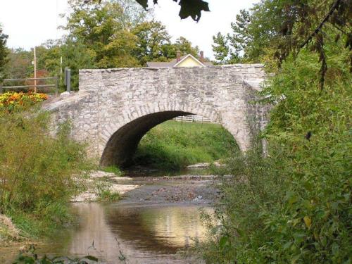 A stone arch bridge spans a calm stream, surrounded by greenery and trees, with a house visible in the background.