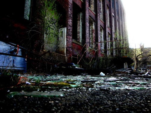Abandoned industrial site with broken glass, overgrown vegetation, and a weathered brick building in the background.