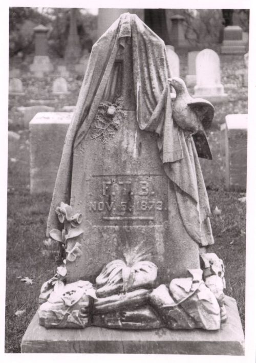 A weathered gravestone draped with a cloth, featuring a dove and floral decorations, set in a cemetery.