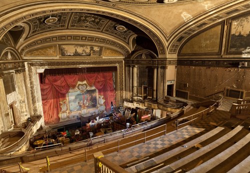 Abandoned theater interior with ornate architecture, a red curtain, and scattered furniture on the stage.