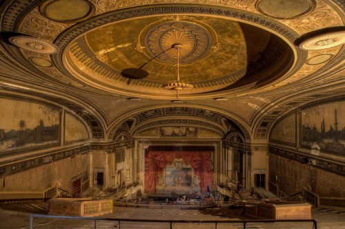 Abandoned theater interior with ornate ceiling, faded murals, and a large stage draped in red curtains. Dusty and neglected.