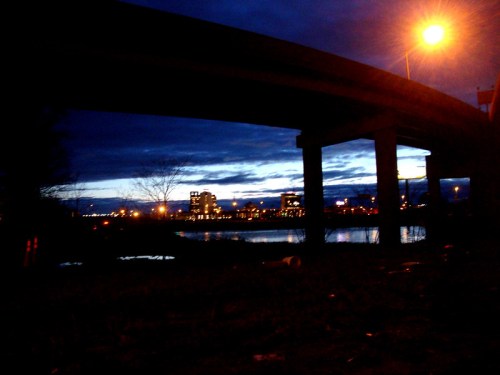 A dark, urban scene under a bridge at dusk, with city lights reflecting on the water and a cloudy sky above.