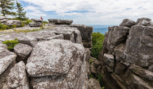 A person stands on rocky cliffs overlooking a lush valley under a cloudy sky.
