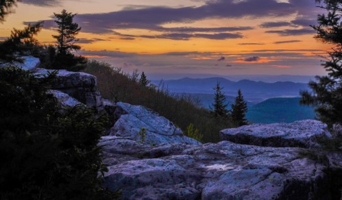 Sunset view from rocky outcrop, with distant mountains and trees silhouetted against a colorful sky.
