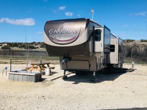 A parked RV labeled "GATEWAY" at a campsite with picnic table and water hookup in a sunny outdoor setting.