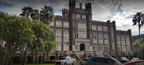 Historic brick building with castle-like architecture, surrounded by palm trees and parked cars under a cloudy sky.