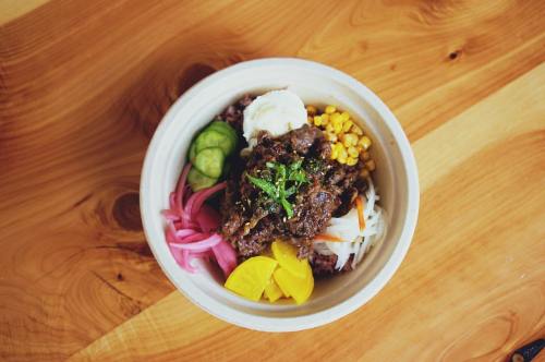 A colorful bowl of rice topped with beef, pickled vegetables, corn, and fresh herbs on a wooden surface.