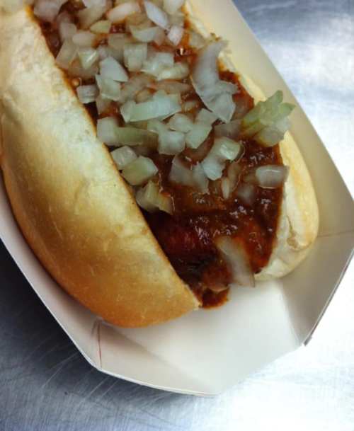 A close-up of a chili dog topped with chopped onions in a white paper tray.