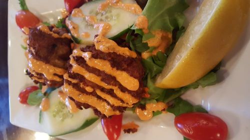 A plate of crispy fritters drizzled with sauce, served on a bed of greens, cherry tomatoes, cucumber, and a lemon wedge.