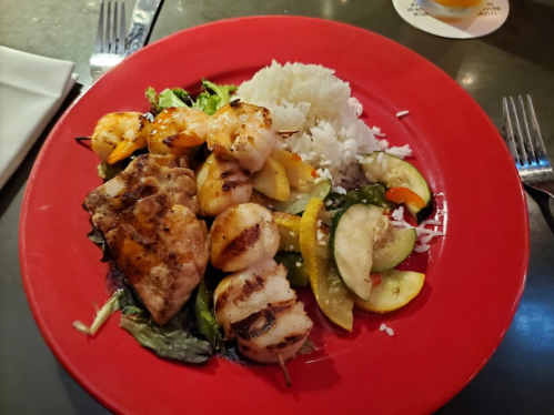 Grilled shrimp and chicken on a bed of greens, served with rice and sautéed vegetables on a red plate.