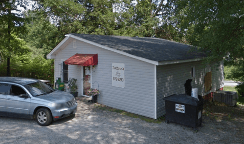 A small gray building with a red awning, parked car, and dumpster nearby, surrounded by trees.