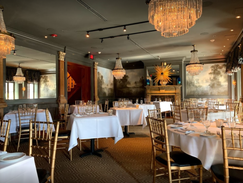 Elegant restaurant interior with white tablecloths, gold chairs, and chandeliers, featuring a warm, inviting atmosphere.