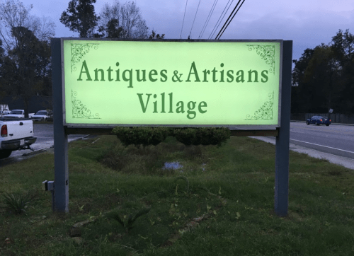 Sign for "Antiques & Artisans Village" illuminated in green, surrounded by grass and trees, near a road.