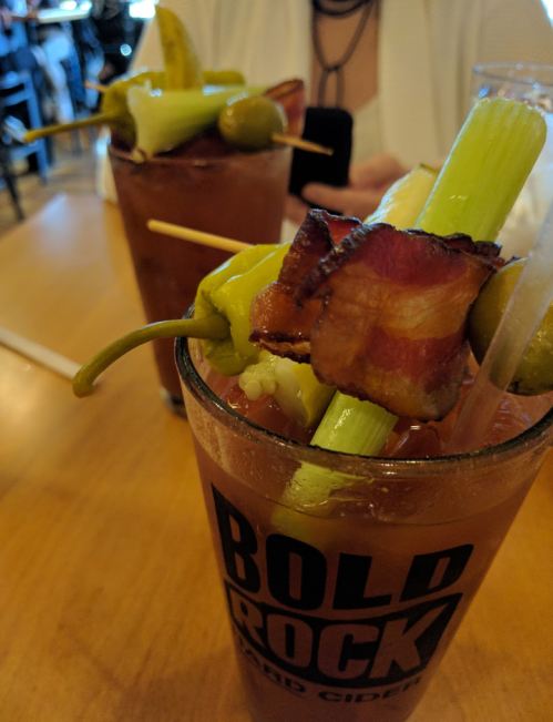 Two cocktails with garnishes of bacon, celery, olives, and peppers, served in glasses on a wooden table.