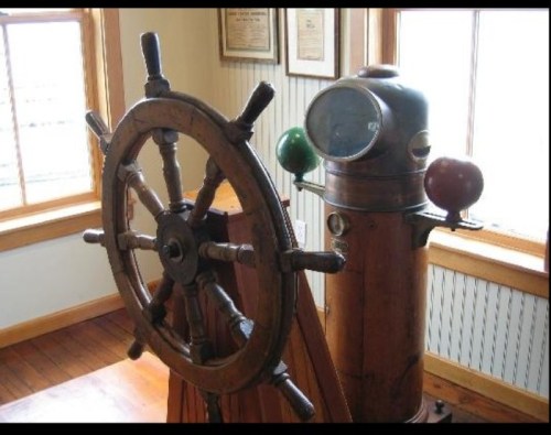 A vintage ship's wheel next to a brass ship's telegraph in a nautical-themed room.