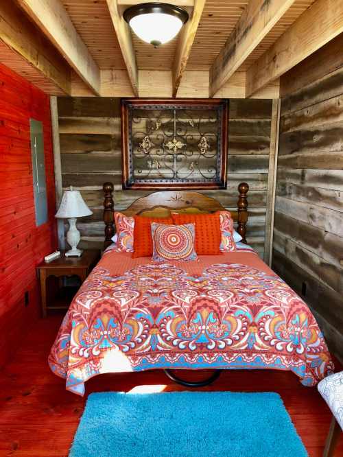 Cozy bedroom with a wooden bed, colorful bedding, and rustic walls, featuring a lamp and decorative wall art.