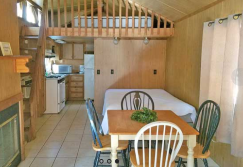 Cozy cabin interior featuring a lofted bed, dining table, and kitchen area with wooden accents and tiled floor.