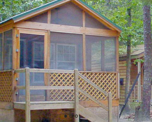 A wooden cabin with a screened porch, surrounded by trees, featuring a staircase and lattice work.