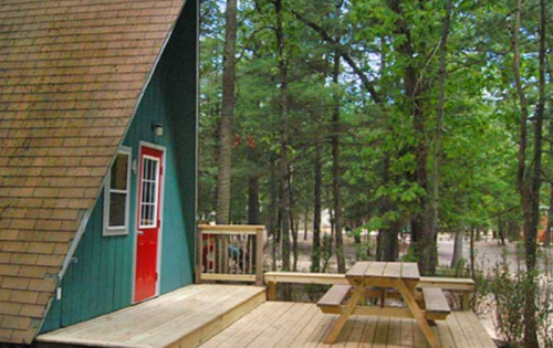 A green A-frame cabin with a red door, surrounded by trees and featuring a wooden deck with a picnic table.