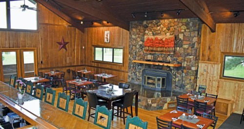 Cozy restaurant interior with wooden walls, stone fireplace, and tables set for dining. Large windows let in natural light.