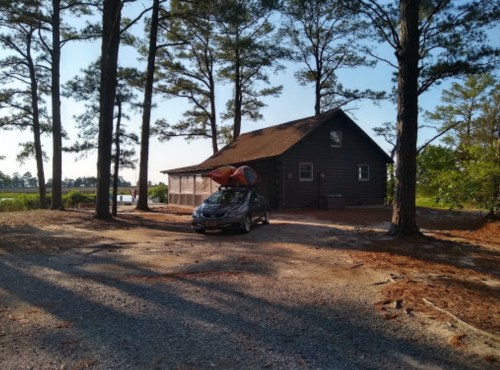 A car with a kayak on top parked near a rustic cabin surrounded by tall trees and a clear sky.