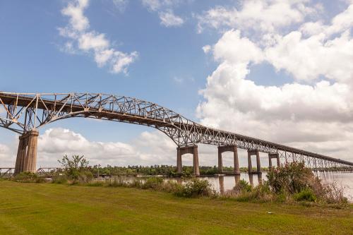 A large steel bridge arches over a river, surrounded by green grass and trees under a partly cloudy sky.