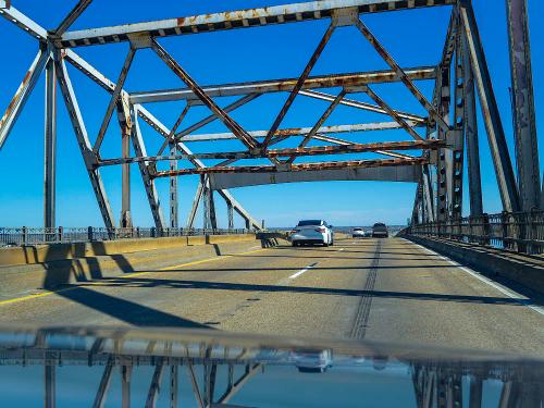 View from a car on a weathered metal bridge, with blue sky and vehicles visible on the road below.