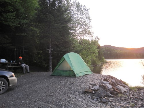 A green tent by a lake at sunset, with a person sitting nearby and a car parked on gravel. Trees surround the area.