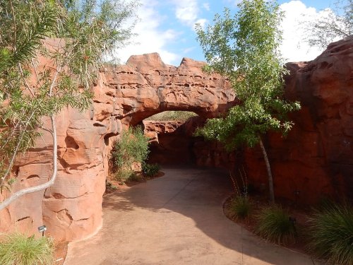 A winding path through red rock formations, with greenery and a natural arch overhead under a blue sky.