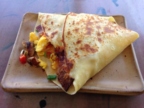 A golden-brown crepe filled with scrambled eggs, vegetables, and meat, served on a brown plate.