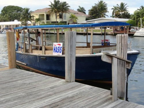 A blue boat docked at a wooden pier, with palm trees and a house in the background. A sign on the boat reads "HASIS."